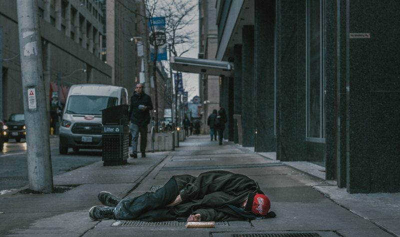 Une personne dort sur le trottoir d'une grande ville. Les passants y sont indifférents.