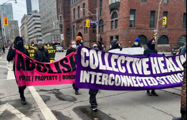 People marching on a street holding pink and purple banners with black and white text.