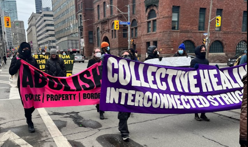 People marching on a street holding pink and purple banners with black and white text.