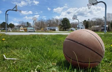 A basketball is seen on the left side of a grassy field with a basketball court and blue sky in the distance.