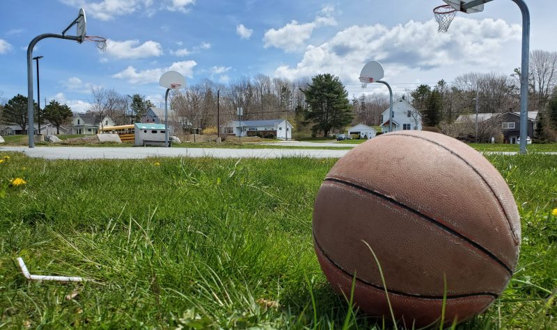 A basketball is seen on the left side of a grassy field with a basketball court and blue sky in the distance.
