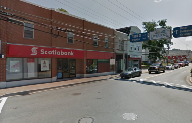 Exterior of a Scotiabank location adjacent to a main street. The building is two stories, made of red brick. There are four small windows on the second story and plate glass windows on the first, with the red Scotiabank banner across the entire building front.