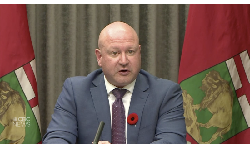 Dr Brent Roussin assit devant deux drapeaux du Manitoba lors d'une conférence de presse.
