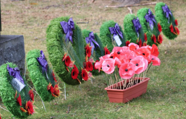 Wreaths are decorated with poppies and purple ribbon. A box of poppies is placed in front of them on the ground.