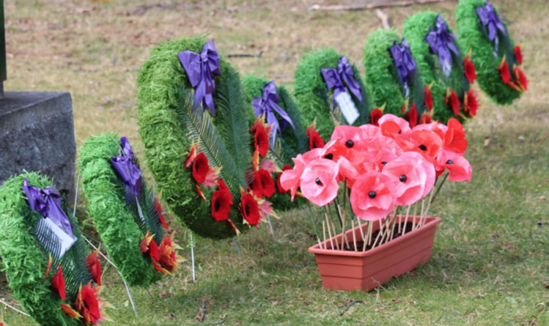 Wreaths are decorated with poppies and purple ribbon. A box of poppies is placed in front of them on the ground.