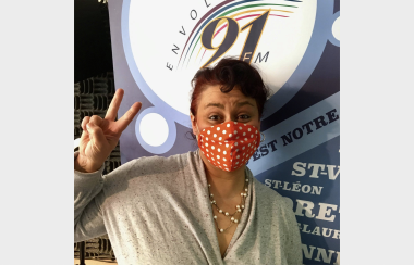 Mélonai est debout devant une affiche d'Envol 91 FM en portant un masque et fait un signe de peace.