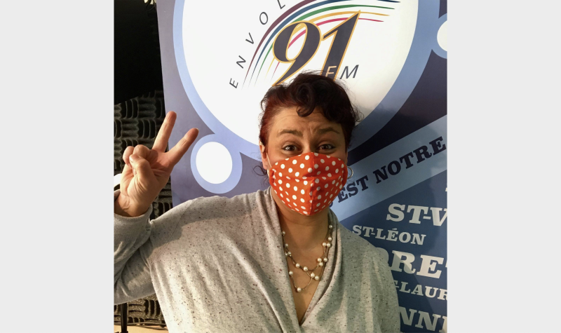 Mélonai est debout devant une affiche d'Envol 91 FM en portant un masque et fait un signe de peace.