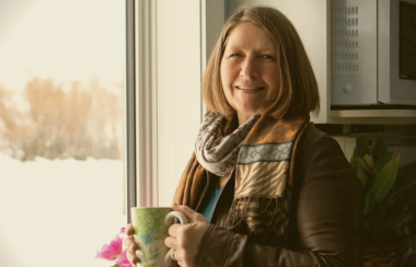 Jeannine est debout devant une fenêtre avec une tasse verte dans les mains.