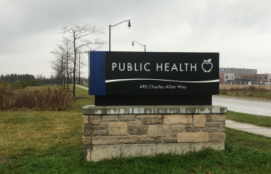 A navy WDG Public Health sign outside out front of their Fergus location on the grass on a cloudy day