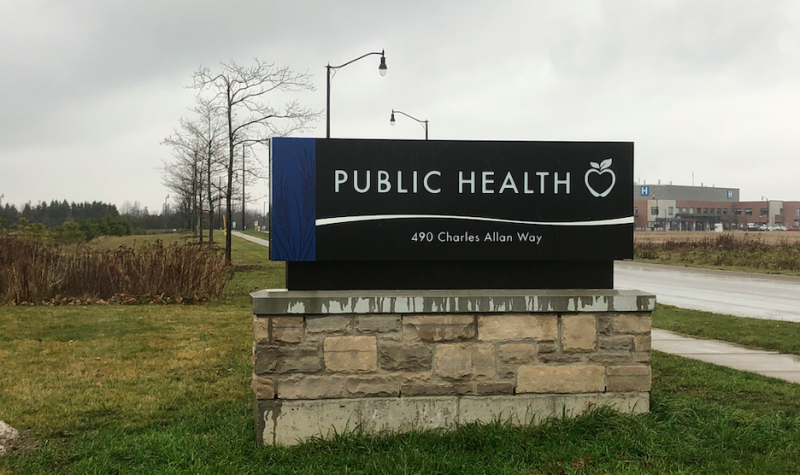 A navy WDG Public Health sign outside out front of their Fergus location on the grass on a cloudy day