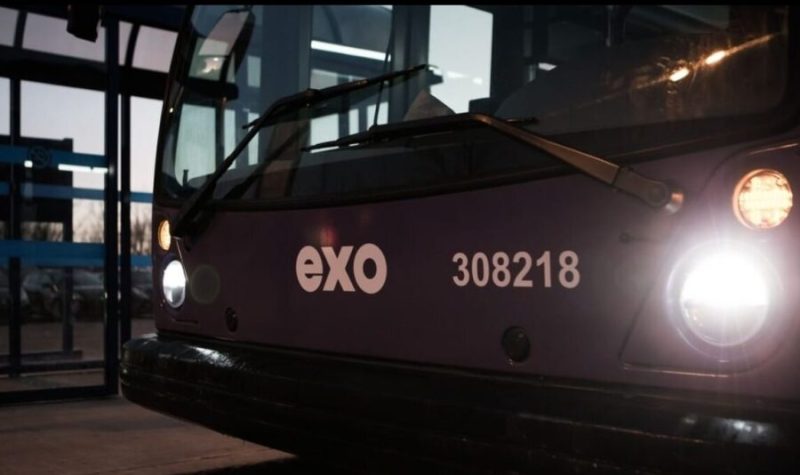 La devanture mauve d'un autobus EXO, les phares allumés, tout près d'un abri-bus au crépuscule.