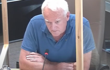 A man leans forward at a council meeting with a microphone in front of him and glass around him