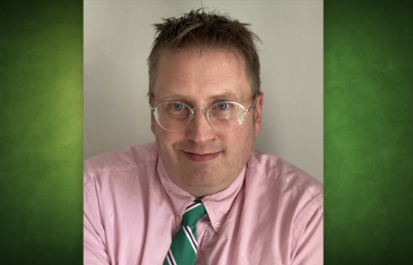 Laurent Poliquin porte des lunettes rondes, une chemise rose et une cravatte verte, il sourit, devant un mur gris.