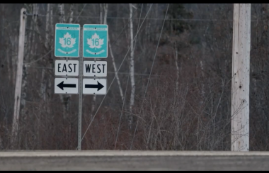 Two green and white signs along highway 16 point east and west witha forest behind