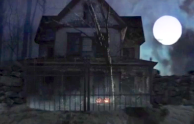 The exterior of an old house with an iron gate around it, pumpkins in the front yard and a full moon in the night sky