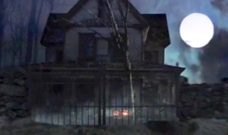 The exterior of an old house with an iron gate around it, pumpkins in the front yard and a full moon in the night sky