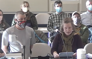 A woman stands at a podium and is speaking, with a man wearing a COVID mask standing beside her and other people sitting in chairs behind them both, listening.