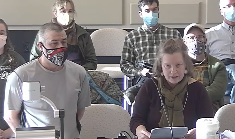 A woman stands at a podium and is speaking, with a man wearing a COVID mask standing beside her and other people sitting in chairs behind them both, listening.