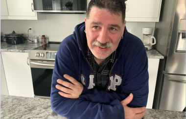 Craig Boutilier is wearing a navy blue sweater and smiling over the table of his apartment kitchen.