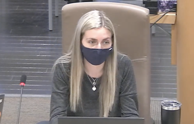 A woman sits at a table with a laptop and a microphone, wearing a mask.