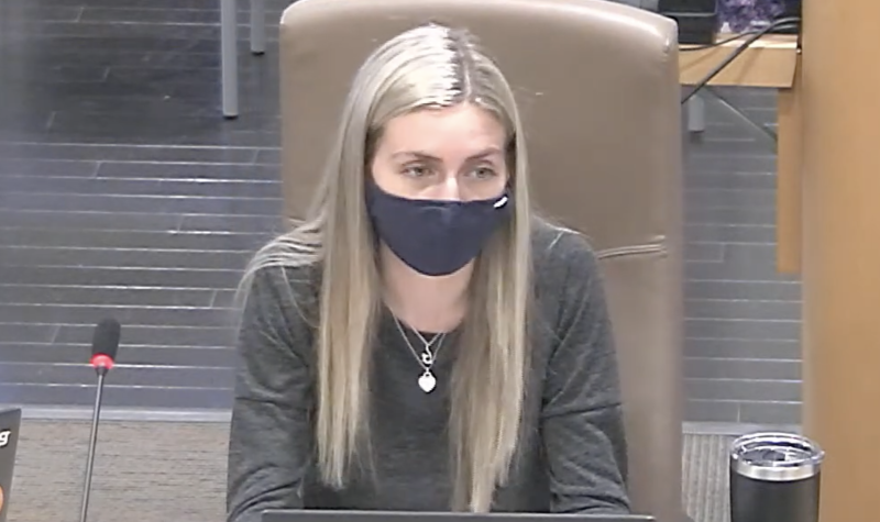 A woman sits at a table with a laptop and a microphone, wearing a mask.