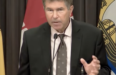 A man in suit and tie standing at a podium speaking, with Canadian and New Brunswick flags in background.