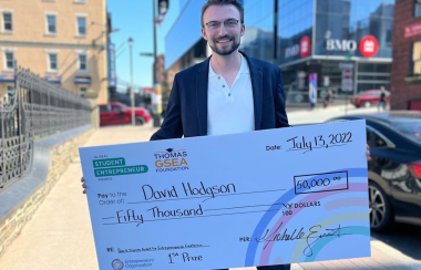 A man wearing a suit holds a $50,000 check from the Global Student Entrepreneur Awards. He stands on the street on a sunny day.