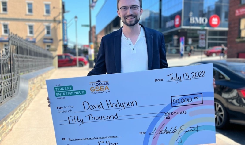 A man wearing a suit holds a $50,000 check from the Global Student Entrepreneur Awards. He stands on the street on a sunny day.