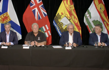 Four men sit side-by-side, dressed in business casual attire, at a black-cloth covered table with microphones, glasses of water and notes. Behind them are the flags representing their respective provinces.