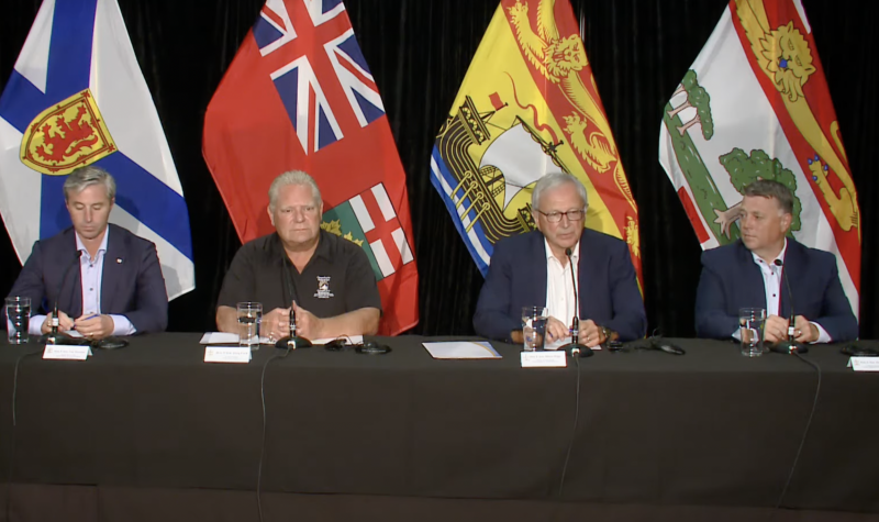 Four men sit side-by-side, dressed in business casual attire, at a black-cloth covered table with microphones, glasses of water and notes. Behind them are the flags representing their respective provinces.