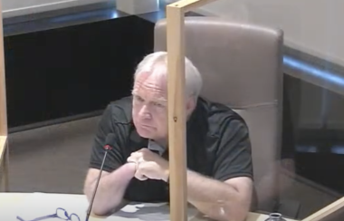 A man with hands folded in front of him sits at a council table with a plexiglass screen beside him, and a microphone in front of him.