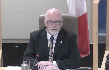 A man in suit and tie sitting at council table with microphone and Canadian flag standing behind him.