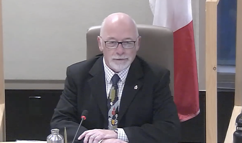 A man in suit and tie sitting at council table with microphone and Canadian flag standing behind him.