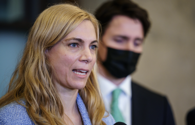 A blonde woman speaks at a microphone. She is wearing a blue sweater. Behind her stands a man wearing a black suit and black mask.