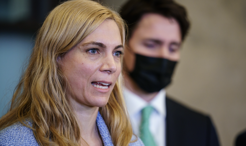 A blonde woman speaks at a microphone. She is wearing a blue sweater. Behind her stands a man wearing a black suit and black mask.