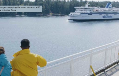 A man wearing yellow and a girl wearing blue stand on a ferry deck looking out at another BC Ferries vessel