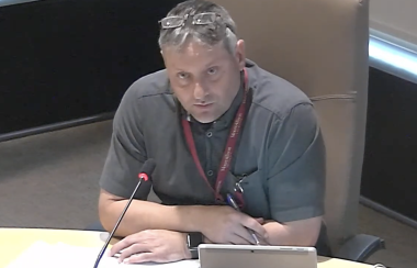 A man sits at a council table with a laptop and microphone.