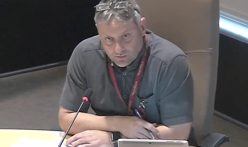 A man sits at a council table with a laptop and microphone.
