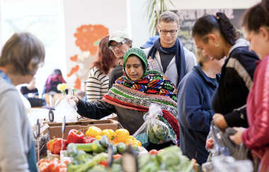 People line up to shop for produce at the North Grover market. Woman in hijab reaches for vegetables, person behind the counter has their back to the camera wearing a grey shirt.