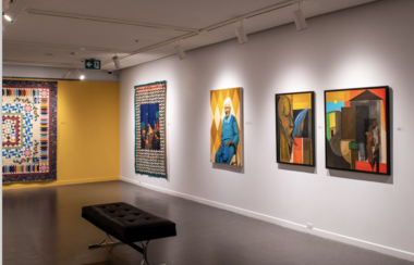 A corner of art gallery. Yellow wall on the left holds a quilt hung on the wall. White wall on the right holds a quilt and three paintings. A bench sits in the middle of the room facing the white wall.