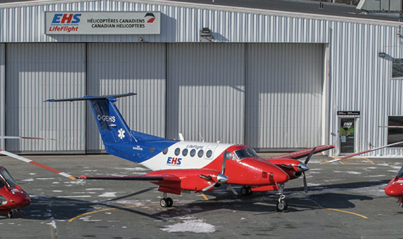 Three EHS LifeFlight aircrafts sit outside of a EHS garage. The two helicopters and plane are blue, white and red stripped.