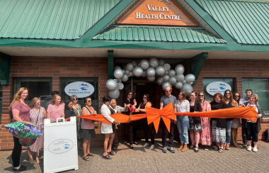 A group of people stand in front of a building called the Valley Heath Centre. They are standing behind an orange ribbon watching as an elderly woman cuts the ribbon with big scissors..