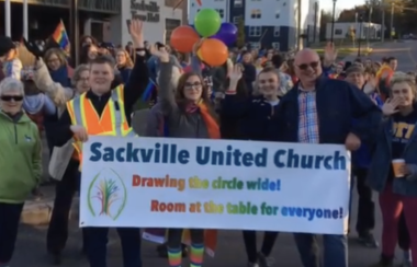 A group of people carrying a rainbow sign reading Sackville United Church. The words on the banner say 'drawing the circle wide'.
