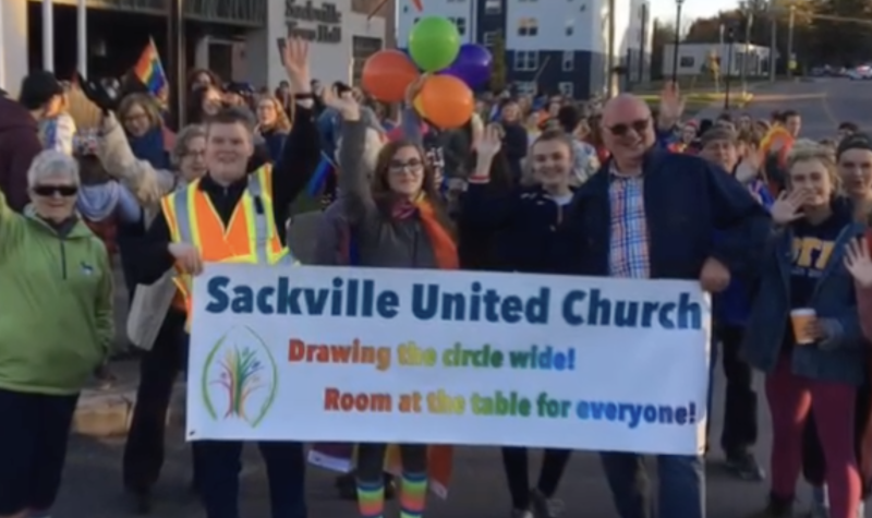 A group of people carrying a rainbow sign reading Sackville United Church. The words on the banner say 'drawing the circle wide'.
