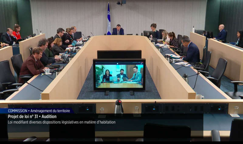 Committee members sit around a room. Three people are presenting remotely from a screen in the middle.