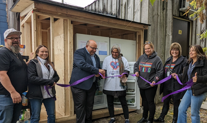 Seven people stand in front of the unfinished community shed building, cutting a purple ribbon.