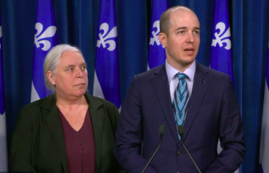 A woman and a man stand behind a podium. Quebec flags are hung in the background.