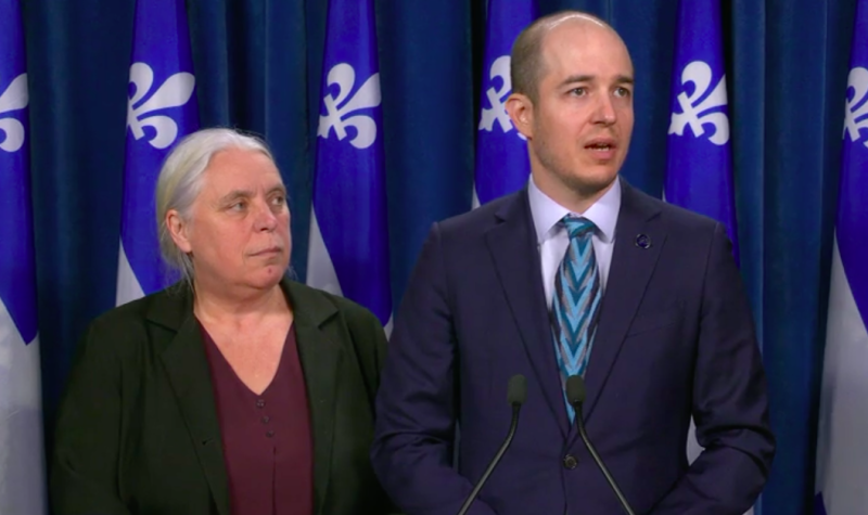 A woman and a man stand behind a podium. Quebec flags are hung in the background.