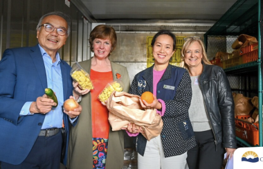 Four people stand in a walk-in refrigerator holding packages of food in their hands, smiling for the camera.