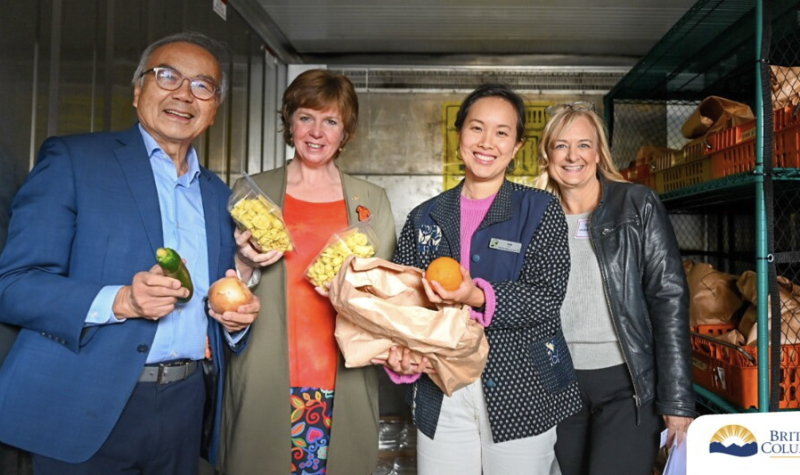Four people stand in a walk-in refrigerator holding packages of food in their hands, smiling for the camera.